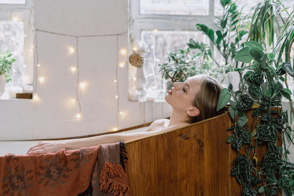 Lady relaxing in a wooden bath surrounded by lush plants and fairy lights