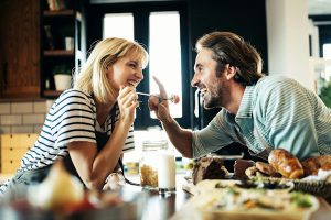 couple in a kitchen she is feeding him with a form he is about to touch her nose with one finger - both smiling