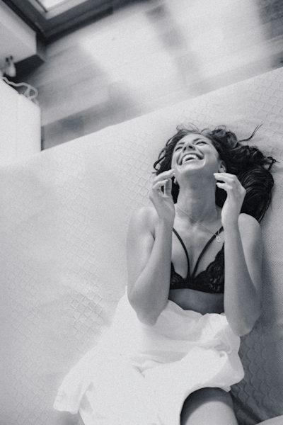 Lady laying on a bed with black bra and arms up to her head she is laughing and covered with a white duvet only