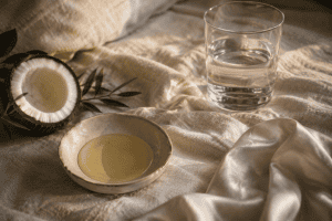 Half a coconut on a bed of linen with silk sheet with small bowl oil & a glass of water